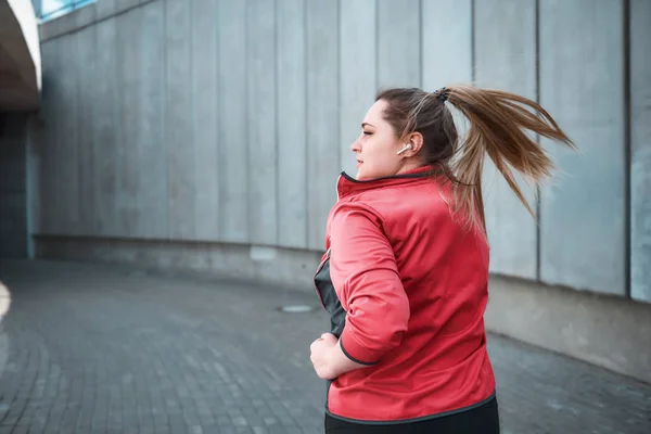 Challenging myself. Back view of young attractive plus size woman in sporty clothes is looking aside and listening music while running in the city