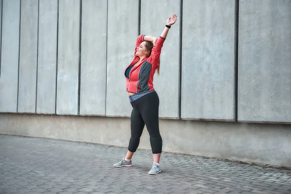 Beautiful plus size woman in sport clothes doing sport outdoors and smiling while standing against gray wall