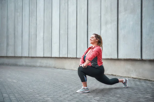 I love sport Happy plus size woman in sport clothes doing stretching exercises and smiling while standing against gray wall.