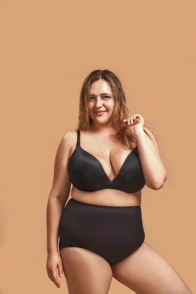 Vertical photo of plus size young woman in sexy black lingerie laughing and posing in studio on brown background