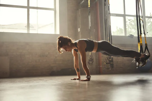 Exercising with suspension straps. Young athletic woman in sports clothing training legs with trx fitness straps in the gym.