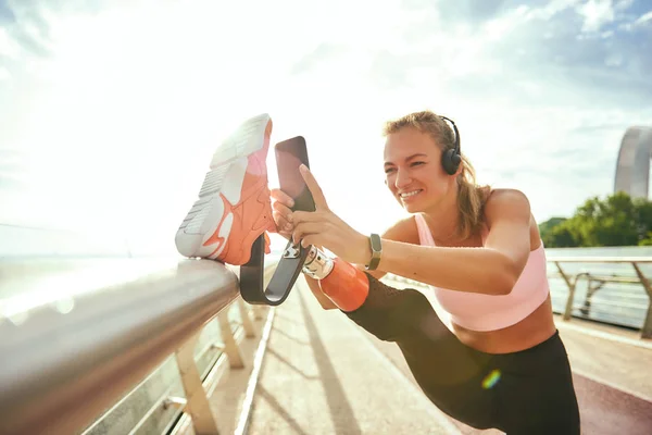 Staying in touch. Happy disabled woman in sportswear and headphones stretching prosthetic leg and looking with smile at her smartphone while standing on the bridge