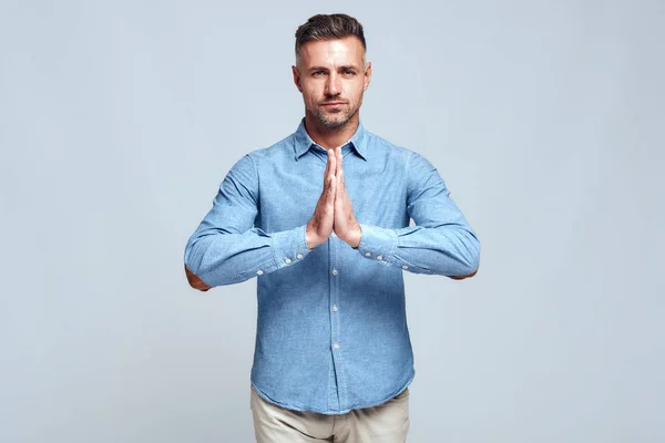 Keep calm. Portrait of young and charming bearded man in casual wear keeping hands clasped and looking at camera while standing against grey background