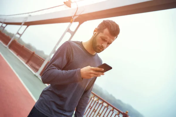Staying in touch. Portrait of young and handsome bearded man in sportswear and headphones using mobile phone while standing outdoors on the bridge in the morning