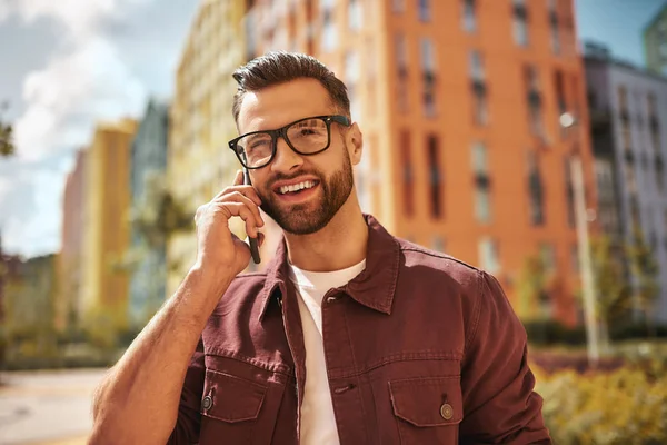 Happy to hear you Portrait of cheerful and handsome bearded man in casual wear and eyeglasses talking by phone and smiling while standing on the street