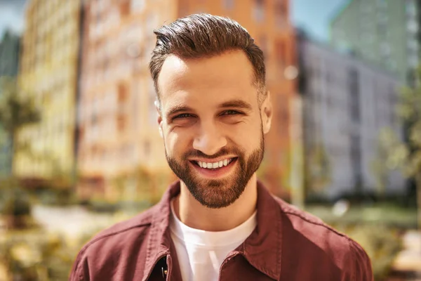 Hello Portrait of happy attractive man with stubble smiling and looking at camera while standing on the street