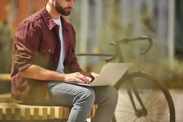 I am always in touch. Side view of young man with stubble in casual clothes working on laptop while sitting on the bench near his bicycle