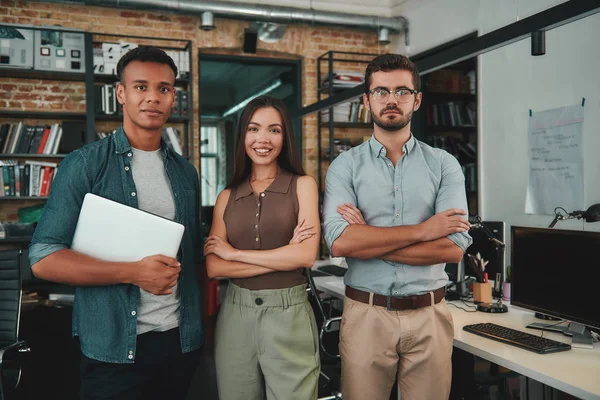 We are perfect team. Group of three cheerful young people in casual wear looking at camera with smile while standing in modern office