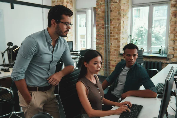 Business experts. Multiethnic group of young employees looking at computer monitor and discussing something while sitting in modern open space
