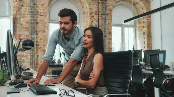 Look here Two young colleagues in casual wear looking at computer monitor and discussing project results while working together in modern office