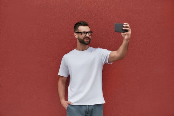 Happy moments. Handsome young man in casual clothes making selfie with his mobile phone and smiling while standing against red wall outdoors