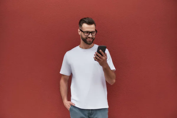 Always in touch. Handsome young man in casual clothes holding mobile phone and smiling while standing against red wall outdoors