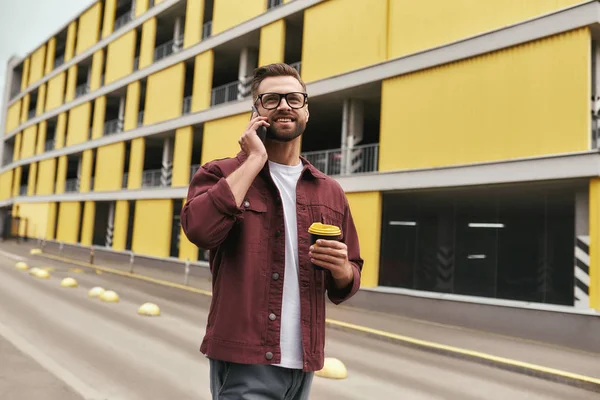 Happy to hear you. Handsome young man in casual wear and eyeglasses holding a disposable cup and talking by phone with somebody while walking through the city street
