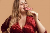 Mmm. Plus size happy woman in velour dress eating red heart-shaped lollipop and keeping eyes closed while standing in studio