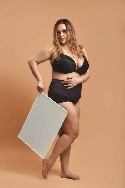 Your text here. Full length of young plus size young woman in black lingerie holding empty blank board and looking at camera while standing against brown background