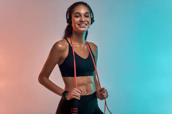 Music is my motivation. Sporty young woman in headphones holding jumping rope on shoulders and looking at camera with smile while standing against colorful background