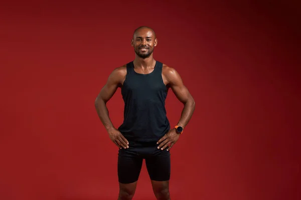 Sport is my life. Handsome and young afro american man in sports clothing keeping arms on hips and looking at camera with smile while standing against red background
