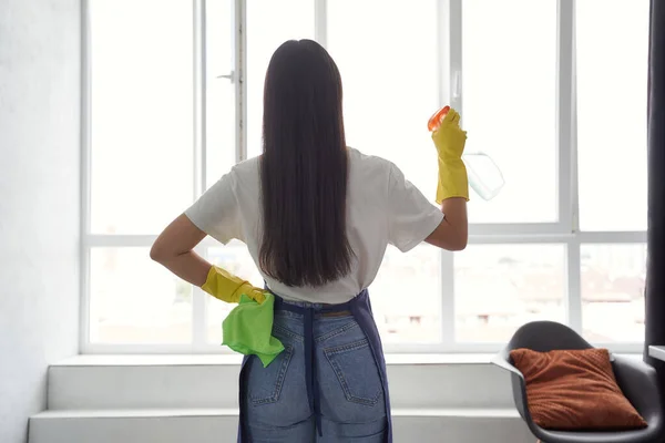 Washing windows professionally. Back view of a young woman, cleaning lady in yellow rubber gloves holding rag and spray while cleaning big window at home