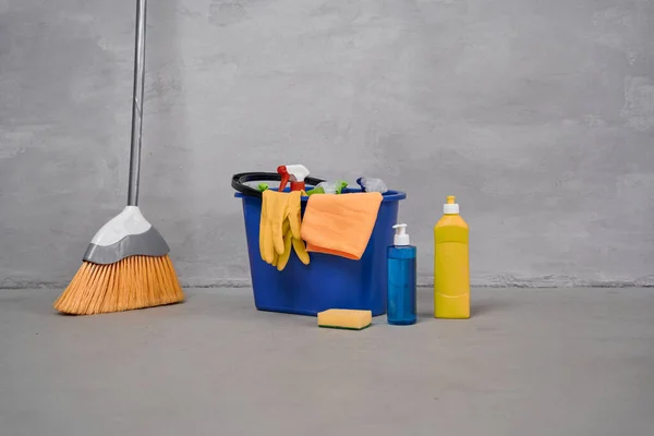 Cleaning supplies. Broom and plastic bucket or basket with cleaning products, bottles with detergents standing on the floor against grey wall
