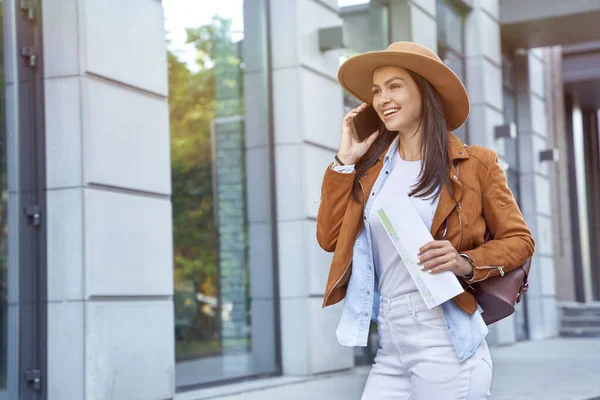 Young and beautiful happy female traveler or tourist wearing hat and backpack holding map and talking by phone with friend or family while walking on city street