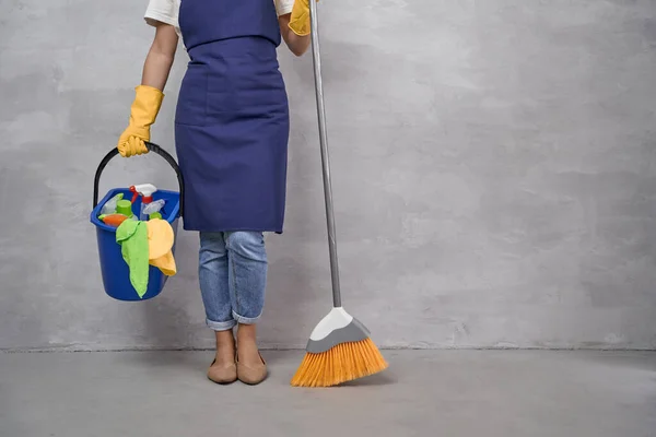 Professional Cleaning Service. Cropped shot of woman in uniform and yellow rubber gloves holding broom and bucket with different cleaning products while standing against grey wall