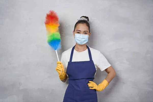 Say No to Dust. Young woman, cleaning lady in uniform wearing medical mask holding colorful microfiber duster and looking at camera while standing against grey wall