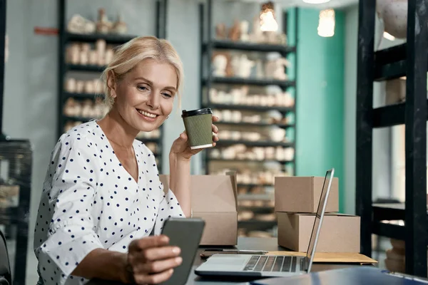 Successful business owner. Beautiful and happy business woman drinking coffee, using smartphone and smiling while working in her art studio or craft pottery shop