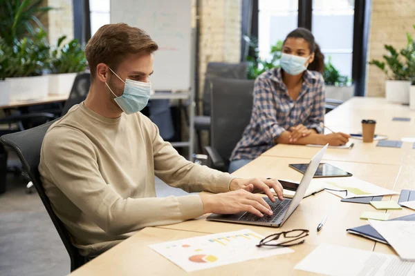 Social distancing concept. Two young colleagues, man and woman wearing medical protective face masks working together in the modern office, guy working on laptop