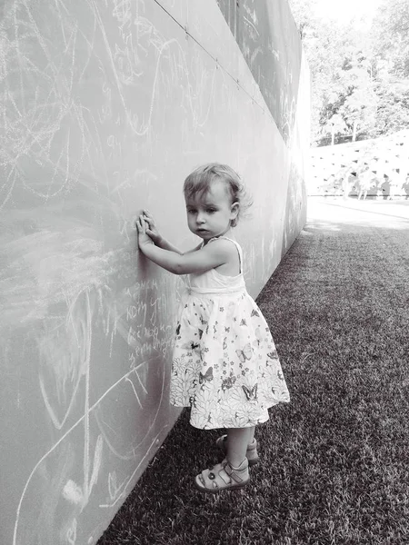 Black and white picture of little girl in a dress standing near the wall