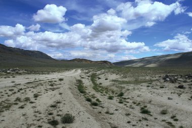 Dağlarda güneşli bir gün, bulutlarla dolu mavi bir gökyüzü, küçük dağlar ve tepeler arasında uzanan bir yol, yakınlarda uzanan taşlar, yolda büyüyen çimenler.