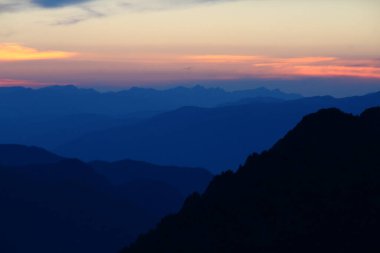 Dağlarda gün batımı, dağların siluetleri bulutlu pembe bir gökyüzüne karşı, dağlarda sis, gölgede yakın bir dağ, mavi ışıkta uzak dağlar