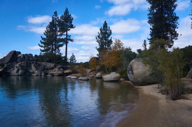 Dev granit kayalar kumlu sahili sonbahar renkleriyle kaplıyor. Arka planda da mavi gökyüzü var.