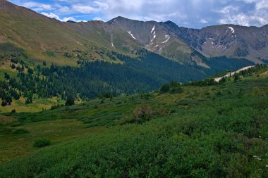 Tepeleri karla kaplı, gökyüzü bulutlu, manzaralı bir dağ çayırı uzanır.