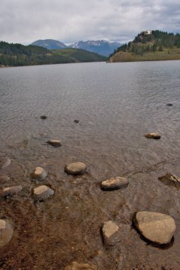 Dağ gölünün ön planındaki kayalar arka plandaki zirvelere kadar uzanıyor.