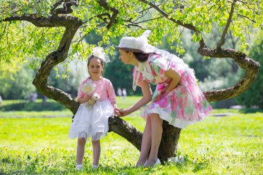 Anne coquettishly gülümseyen bir çocuk elinden tutar. Bayanlar vintage kıyafetler sıradışı bir ağacın altında oturup. Beyaz pembe yeşil elbise ve şapka. Anne ve kızı bir yaz parkta.