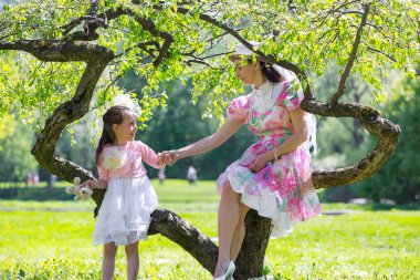 Anne coquettishly gülümseyen bir çocuk elinden tutar. Bayanlar vintage kıyafetler sıradışı bir ağacın altında oturup. Beyaz pembe yeşil elbise ve şapka. Anne ve kızı bir yaz parkta.