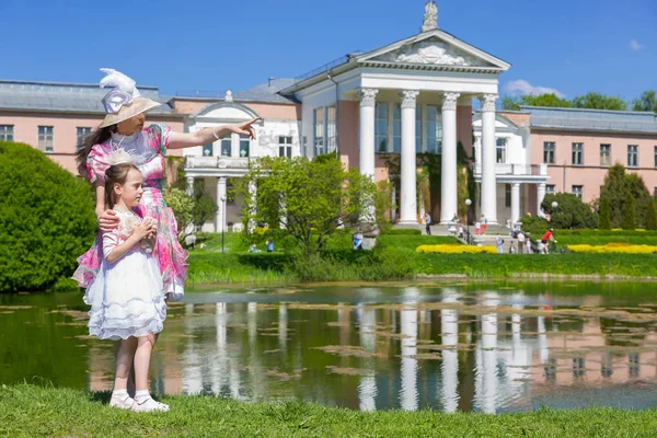 Ladies in vintage clothes stand near the lake against the backdrop of the historic palace. White pink green dress, a hat and beads made from imitation jewelry. Mother and daughter in a summer park.