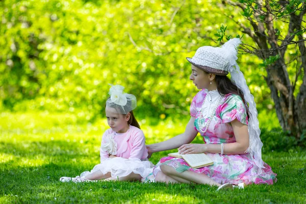 Aile İdil, güzel ışık vintage elbiseler boncuk ve kitap okuma ve birlikte dalga geçmeyi şapkalar, güneşli bir parlak parkta yeşil çimenlerin üzerinde oturuyor. Anne ve kızı bir yaz parkta.