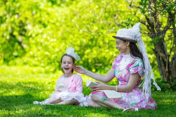家庭田园风光 坐在绿色的草地上 在阳光明媚的公园里 在美丽的灯光下 珠子和帽子 看书和鬼混在一起 母亲和女儿在一个夏日公园里 — 图库照片