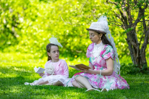 Family idyll, sitting on green grass in a sunny bright park in beautiful light vintage dresses in beads and hats, reading books and fooling around together. Mother and daughter in a summer park.