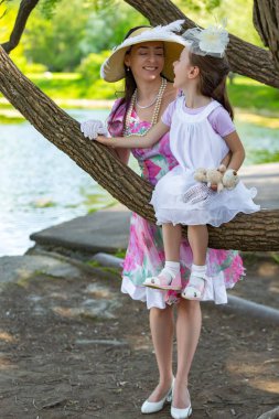 Bir ağaç dalı, gülümsüyor ve gülüyor, beyaz bir ışık elbiseli kızı oturur. Güzel bir şapka ve eldiven hugs yanında anne stand. Anne aşk portresi. Aile bir yaz parkta.