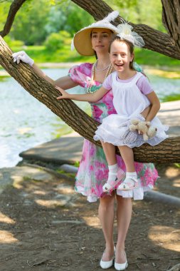 Bir ağaç dalı, gülümsüyor ve gülüyor, beyaz bir ışık elbiseli kızı oturur. Güzel bir şapka ve eldiven hugs yanında anne stand. Anne aşk portresi. Aile bir yaz parkta.