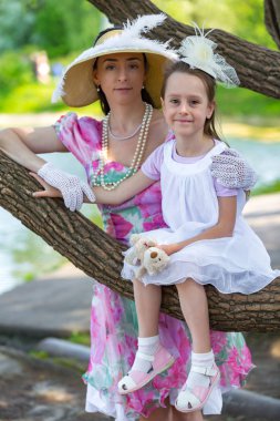 Bir ağaç dalı, gülümsüyor ve gülüyor, beyaz bir ışık elbiseli kızı oturur. Güzel bir şapka ve eldiven hugs yanında anne stand. Anne aşk portresi. Aile bir yaz parkta.