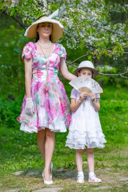 Anne ve kızı bahar ormanda çiçek açması bir elma ağacı arka planı poz. Onlar ışık tarihsel elbiseler, şapka ve ellerinde bir fan ile stand. Aile bir yaz parkta.