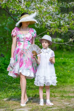 Anne ve kızı bahar ormanda çiçek açması bir elma ağacı arka planı poz. Onlar ışık tarihsel elbiseler, şapka ve ellerinde bir fan ile stand. Aile bir yaz parkta.