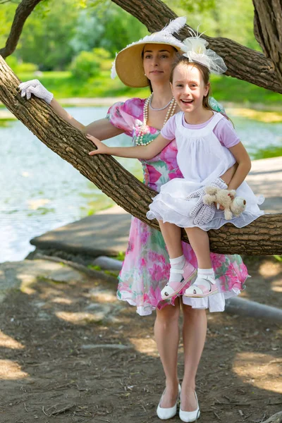 Bir ağaç dalı, gülümsüyor ve gülüyor, beyaz bir ışık elbiseli kızı oturur. Güzel bir şapka ve eldiven hugs yanında anne stand. Anne aşk portresi. Aile bir yaz parkta.