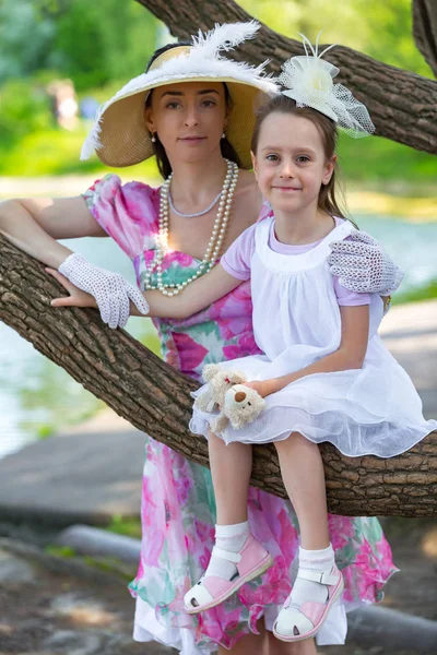 Bir ağaç dalı, gülümsüyor ve gülüyor, beyaz bir ışık elbiseli kızı oturur. Güzel bir şapka ve eldiven hugs yanında anne stand. Anne aşk portresi. Aile bir yaz parkta.