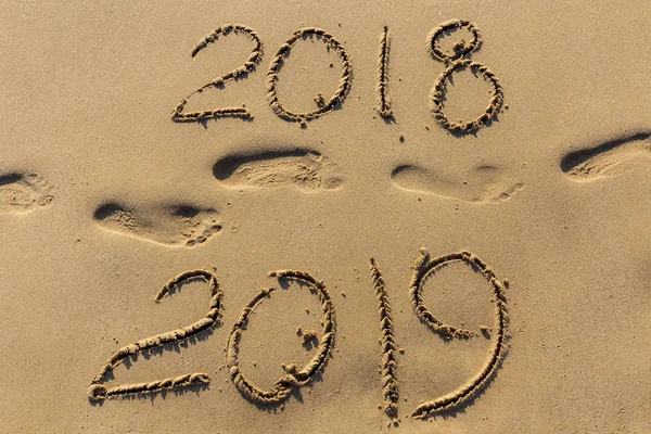 Concept of Happy New Year 2019 is coming and leaving year of 2018. Text on the sea beach and footprints on the sand. Wave water covering digits inscription. Meet at a tropical resort in Thailand.