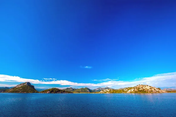 美丽光滑的蓝色湖面映出了蓝天与白云 丘陵地形与树木在岸边的石坡 黑山波德戈里察地区斯卡达尔湖 — 图库照片