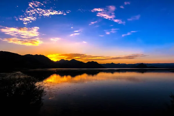 傍晚湖面上的五颜六色的日落 橙色的黄色太阳落在美丽的蓝天下的黑色山脉的轮廓后面 黑山波德戈里察地区斯卡达尔湖 — 图库照片